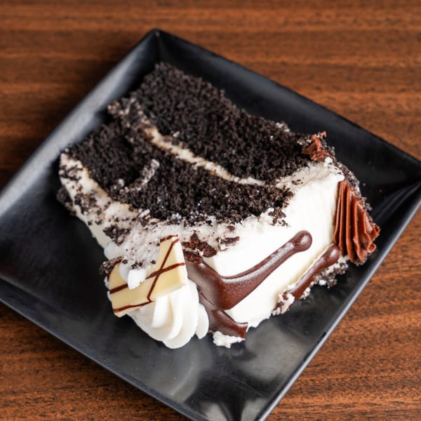 A slice of chocolate cake with white frosting, chocolate drizzle, and chocolate shavings on a black plate.